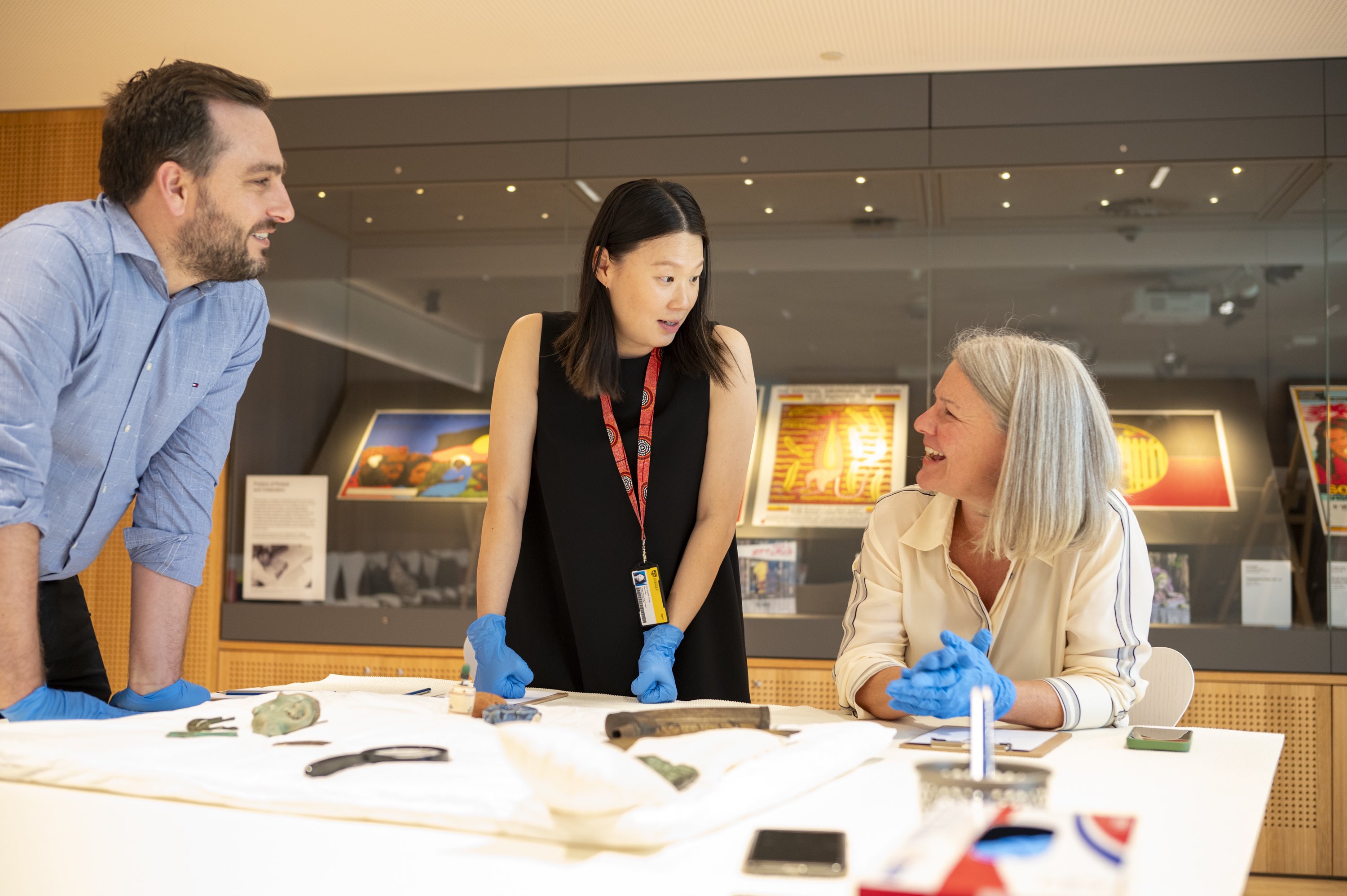 Three people sit at a table wearing latex gloves for handling special objects while talking and laughing